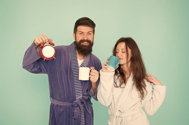 Couple in bathrobes with mugs. Man with beard and sleepy woman enjoy morning coffee or tea. Time to wake up and have nice day. Guy husband hold coffee and clock. Every morning begins with coffee - Foto, imagen