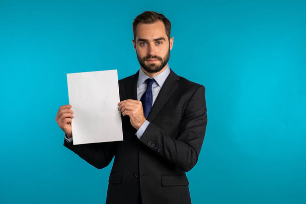 Businessman holding white vertical a4 paper poster. Copy space. Smiling handsome man in corporate suit with tie on blue background. - Foto, afbeelding