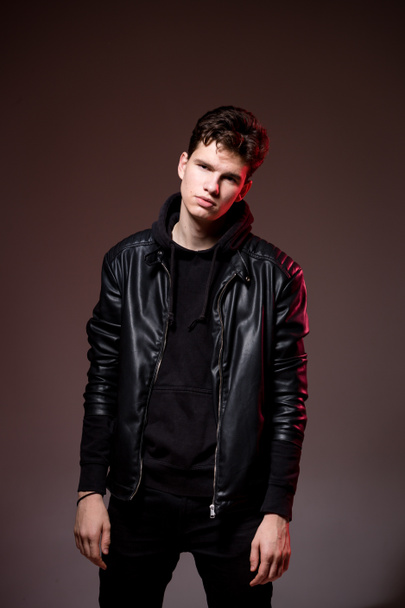 Attractive young guy posing in studio. Young caucasian guy model in black clothes posing in the studio on a dark burgundy background with red backlight. Man in a leather jacket - Foto, Bild