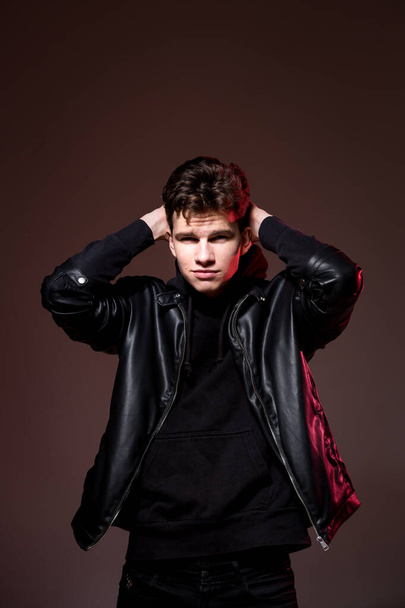 Attractive young guy posing in studio. Young caucasian guy model in black clothes posing in the studio on a dark burgundy background with red backlight. Man in a leather jacket - Foto, afbeelding