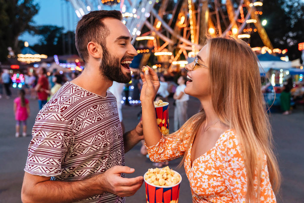 Positive loving couple in amusement park eat popcorn. - Фото, изображение