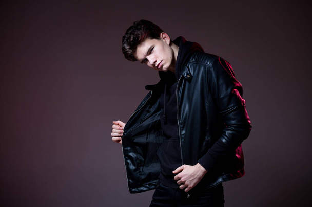 Attractive young guy posing in studio. Young caucasian guy model in black clothes posing in the studio on a dark burgundy background with red backlight. Man in a leather jacket - Fotoğraf, Görsel