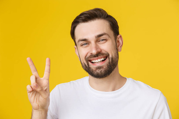 Jonge knappe man draagt gestreept t-shirt over geïsoleerde gele achtergrond glimlachend naar de camera kijkend met vingers die een overwinningsbord doen. Nummer twee - Foto, afbeelding