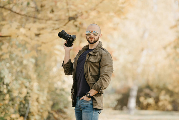 Fotógrafo careca com barba em óculos de sol aviador com lentes espelhadas, jaqueta militar de carga de azeitona, jeans azuis e camisa com relógio de pulso digital segura a câmera em uma mão dobrada na floresta
. - Foto, Imagem