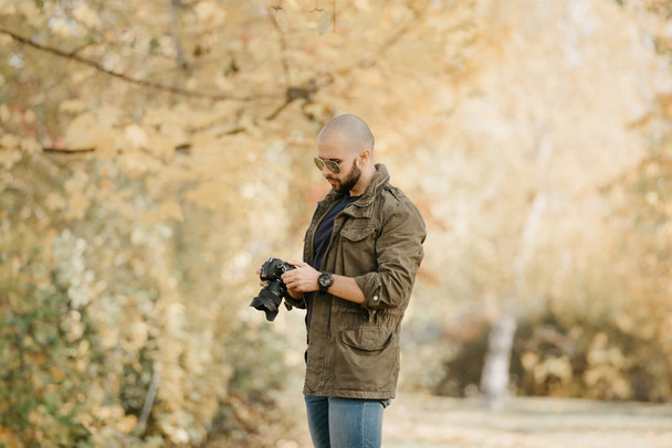 Bald photographer with a beard in aviator sunglasses with mirror lenses, olive cargo military jacket, blue jeans and shirt with digital wristwatch scrolls photos on the camera in the forest. - Foto, Imagem