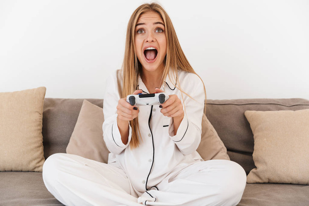 Photo of excited blonde woman playing video game while sitting on sofa - Foto, Bild