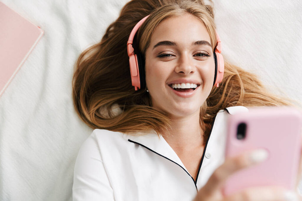Image of girl in headphones holding cellphone while lying in bed - Foto, Imagem