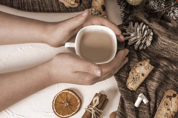 Handen van een man met een kop koffie met melk op een tafel met traditionele Italiaanse koekjes Biscotti met hazelnoten, amandelen, walnoten op een grijze achtergrond met schijfjes gedroogde sinaasappelen en kaneel. - Foto, afbeelding