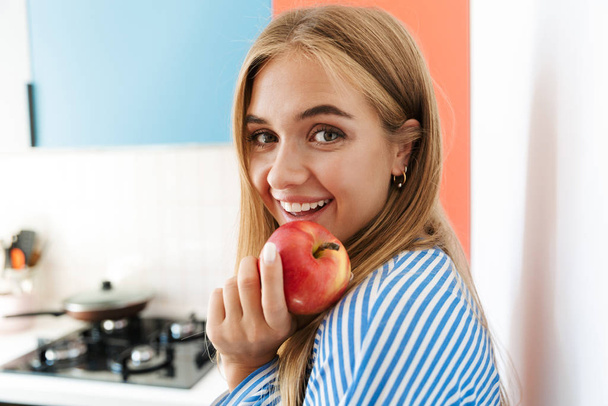 Image of joyous young girl smiling and eating apple in kitchen - Valokuva, kuva