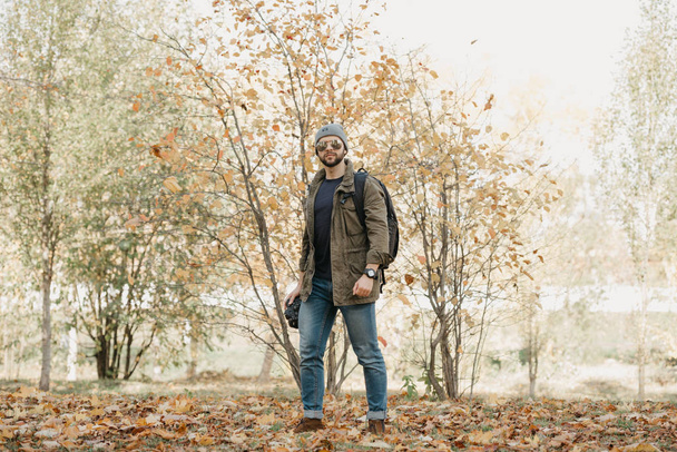 Военный храбрый журналист с бородой в летных солнцезащитных очках с зеркальными линзами, оливковой боевой курткой, джинсами и рубашкой с наручными часами держит камеру DSLR, смотрит вокруг леса в полдень
 - Фото, изображение