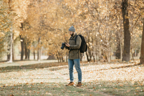 A traveler with a beard in an olive military cargo combat jacket, jeans, hat with backpack and wristwatch holds the DSLR camera and checks something in the smartphone in the forest at the noon. - Photo, Image