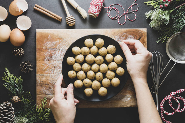 Christmas cookies. Top view of female hands holding, cooking raw unbaked dough balls in black plate with Christmas decoration. - Foto, imagen