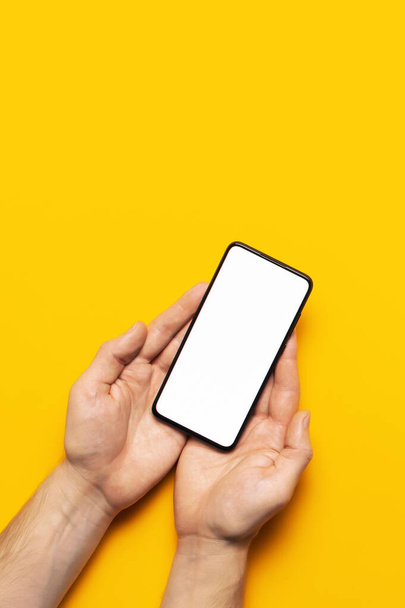 Las manos masculinas sostienen un teléfono inteligente negro moderno con pantalla blanca en blanco sobre fondo amarillo. Tecnología moderna, teléfono, gadget en manos, pantalla táctil, plantilla para su diseño. Burla.
 - Foto, imagen