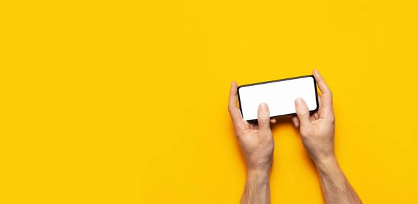 Les mains masculines tiennent un smartphone noir moderne avec écran blanc sur fond jaune. Technologie moderne, téléphone, gadget dans les mains, écran tactile, modèle pour votre conception. Maquette
 - Photo, image