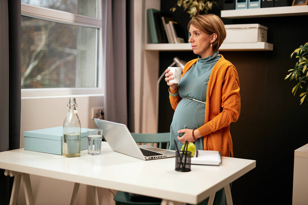 Uroczy biały w ciąży bizneswoman stoi w biurze przy biurku, trzymając kubek z herbatą, dotykając brzucha i patrząc przez okno. - Zdjęcie, obraz