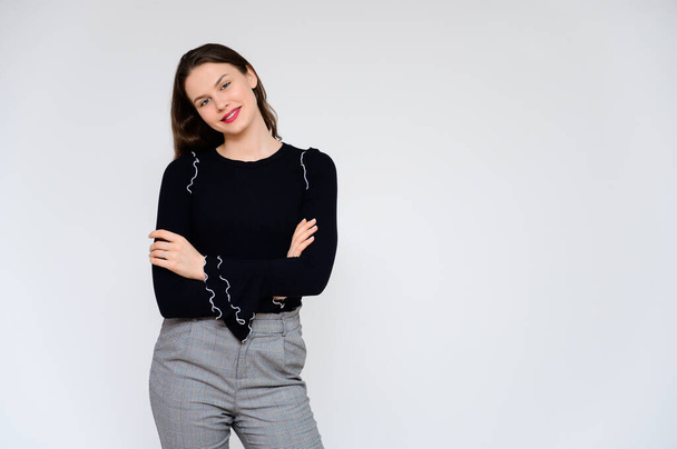 Concept adult girl on a white background. A photo of a pretty brunette girl in gray trousers and a black sweater smiles and shows different emotions in different poses right in front of the camera. - Foto, afbeelding