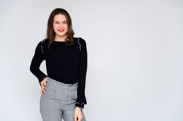 Concept adult girl on a white background. A photo of a pretty brunette girl in gray trousers and a black sweater smiles and shows different emotions in different poses right in front of the camera. - Fotó, kép