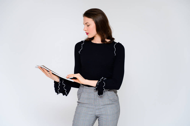 Concept adult girl on a white background with a white folder in hands. A photo of a pretty brunette girl in gray trousers and a black sweater smiles and shows different emotions in different poses. - Fotoğraf, Görsel