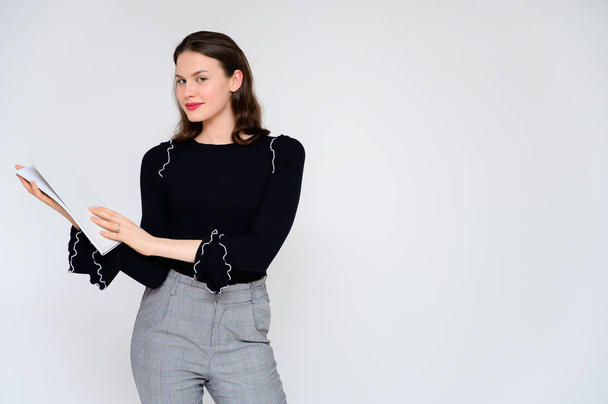 Concept adult girl on a white background with a white folder in hands. A photo of a pretty brunette girl in gray trousers and a black sweater smiles and shows different emotions in different poses. - Photo, Image