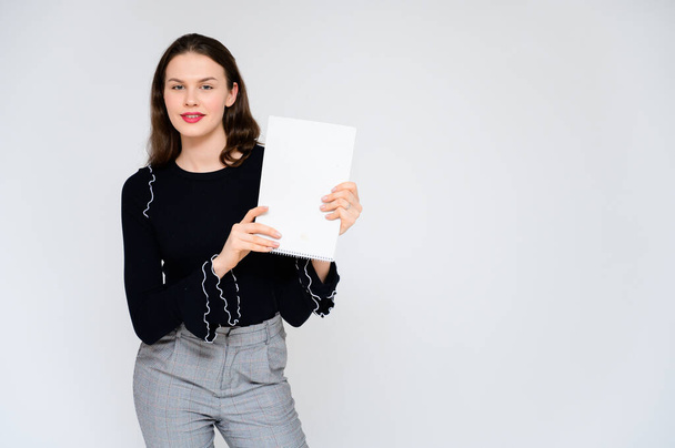 Concept adult girl on a white background with a white folder in hands. A photo of a pretty brunette girl in gray trousers and a black sweater smiles and shows different emotions in different poses. - 写真・画像