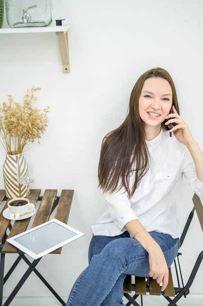 A woman in a white shirt and jeans sits in a cafe and drinks coffee. Female office worker at the lunch break talking on the phone. Manager at a business meeting in a restaurant - Фото, изображение