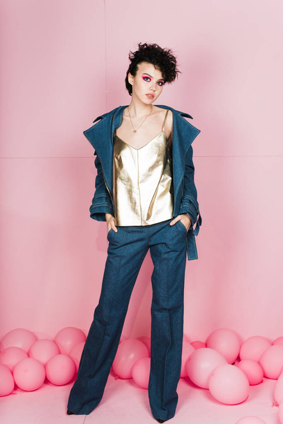 A girl with a short cool flying hairstyle in a denim suit and a gold T-shirt on a pink pastel background. Portrait of a cheeky model in the studio - Photo, Image