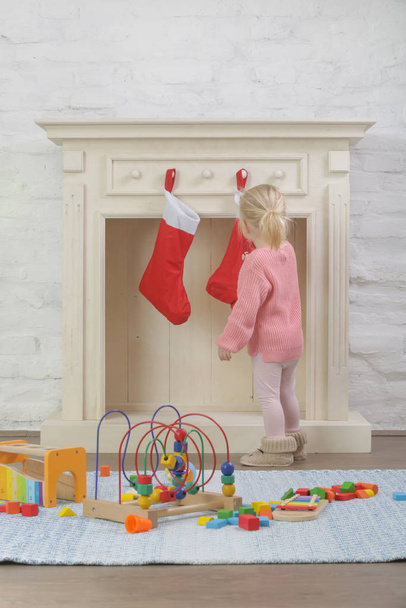Adorable toddler girl playing indoor in front of wooden fireplace decorated with red Christmas socks, winter holidays at home - Photo, Image
