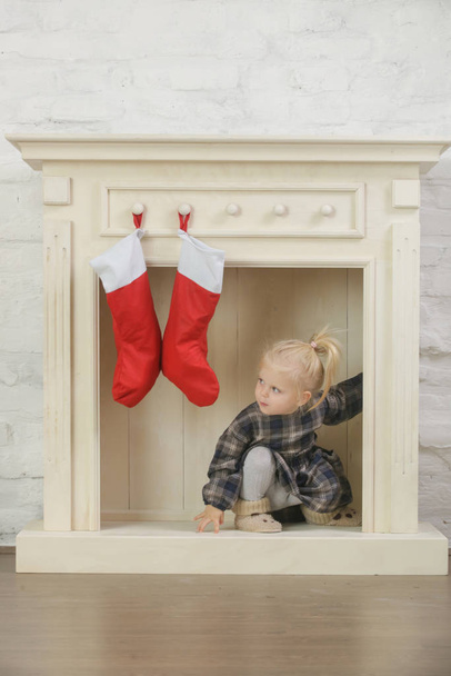 Adorable toddler girl playing indoor in front of wooden fireplace decorated with red Christmas socks, winter holidays at home - Foto, Imagem