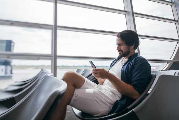 Een man zit in een wachtkamer bij de gate op het vliegveld met een telefoon. - Foto, afbeelding