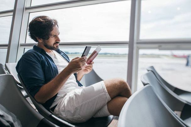 Un hombre se sienta en una sala de espera cerca de la puerta en el aeropuerto con un teléfono
. - Foto, imagen