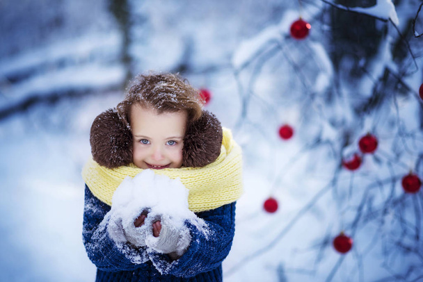 Little smiling girl in a blue pullover, fluffy brown headphones and a yellow scarf with snow in her hands. Winter vacations, winter play, joy and happines. Outdoor winter - Photo, Image