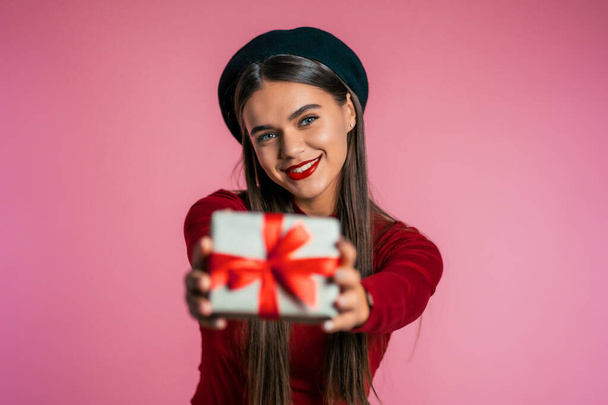 Pretty woman gives gift and hands it to the camera. She is happy, smiling. Girl on pink background. Positive holiday shot.  - Foto, Imagen