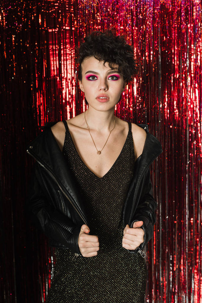 A lovely young girl in a dress and leather jacket is standing on a silver red background made of rain, tinsel. A model with a short stylish flying hairstyle on a shiny Christmas background with bright - Foto, Bild