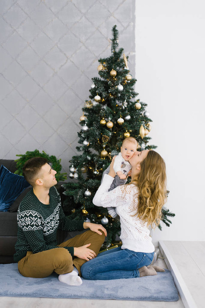 クリスマスと新年明けましておめでとうございます。若い家族によるお祝い。クリスマスツリーの近くに座っている親と赤ちゃん。ママキス赤ちゃん - 写真・画像
