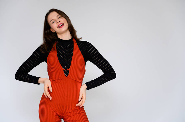 Concept adult girl on a white background. A photo of a pretty brunette girl in red trousers and a black sweater smiles and shows different emotions in different poses right in front of the camera. - Fotó, kép