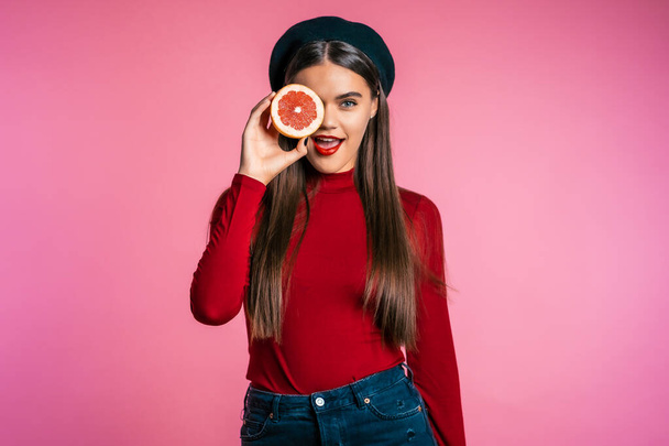 Portrait of young beautiful woman in red with two half of juicy grapefruit on pink studio background. Healthy eating, dieting, antioxidants concept. - Фото, изображение