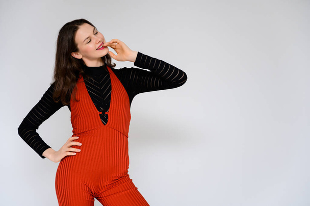 Concept adult girl on a white background. A photo of a pretty brunette girl in red trousers and a black sweater smiles and shows different emotions in different poses right in front of the camera. - Foto, afbeelding