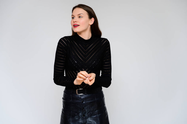 Concept adult girl on a white background. A photo of a pretty brunette girl in a black skirt and black sweater smiles and shows different emotions in different poses right in front of the camera. - Foto, imagen