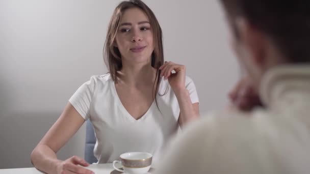 Face of charming Caucasian young woman talking emotionally to unrecognizable lady at the foreground. Brunette daughter telling mom about news in her life. Women resting and drinking hot tea at home. - Video