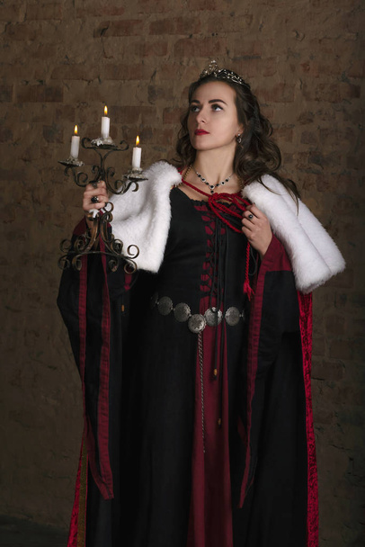 Beautiful young woman in medieval dress with crown at the wall background. Fairytale fantasy scene. Dungeon - Photo, Image