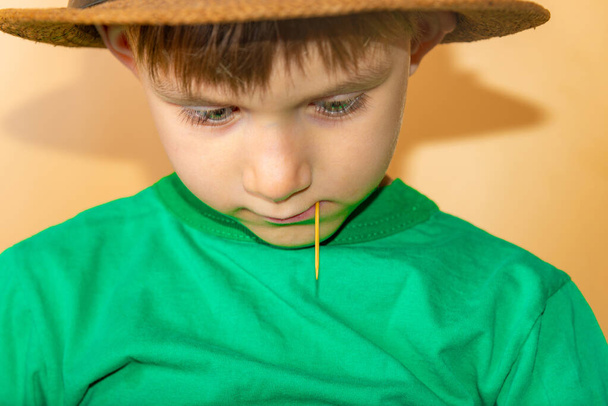 Ένα αγόρι με ένα ψάθινο καπέλο με μια οδοντογλυφίδα στα δόντια του κοιτάει κάτω. - Φωτογραφία, εικόνα