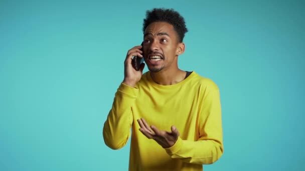 Angry african manin yellow wear screaming down, swears with somebody. Using mobile phone. Stressed and depressed man on blue background. - Footage, Video