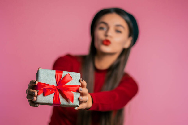 Pretty woman gives gift and hands it to the camera. She is happy, smiling. Girl on pink background. Positive holiday shot.  - Photo, image