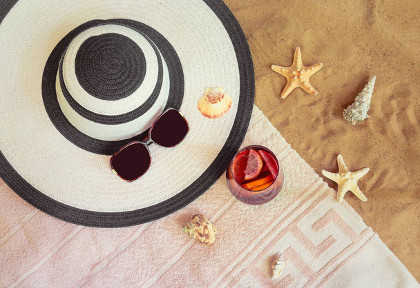 Large beach hat, sunglasses, glass of sangria, starfishes, seashells  and towel on sand beach.  - Фото, изображение
