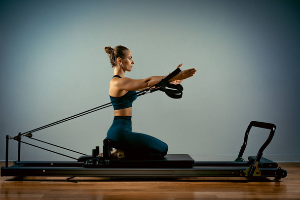 Young girl doing pilates exercises with a reformer bed. Beautiful slim fitness trainer on reformer gray background, low key, art light. Fitness concept - Foto, immagini