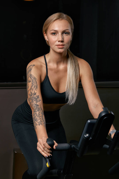 Young confident woman athlete exercising on bicycle indoors. Attractive determined fitness girl doing cycling exercises in dark gym. Functional workout training of sports girl. Cardio training - Fotoğraf, Görsel