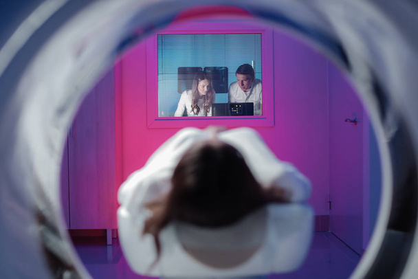 Kız hasta tomografide yatıyor ve tarama için bekliyor. Muayene odasından üç doktor resimlere baktı. - Fotoğraf, Görsel