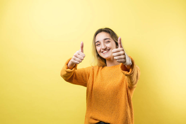 Happy young caucasian female in an orange sweater making thumb up sign and smiling. Good job and respect. - Photo, Image