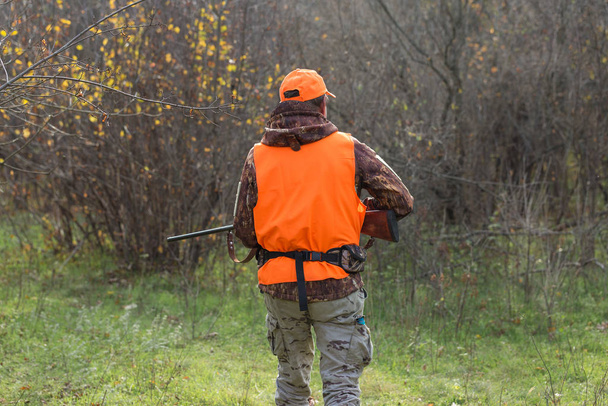 A man with a gun in his hands and an orange vest on a pheasant hunt in a wooded area in cloudy weather. Hunter with dogs in search of game. - Foto, Imagem