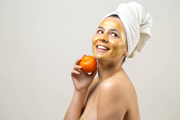 Piękny portret kobiety w białym ręczniku na głowie ze złotą maską odżywczą na twarzy. Oczyszczanie skóry ekologiczny kosmetyczny spa zrelaksować koncepcję. Dziewczyna stoi z tyłu trzymając pomarańczowy mandarynkę. - Zdjęcie, obraz